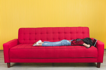 Studio shot Asian young primary school girl in denim jeans overalls outfit laying down sweet dream sleeping on red cozy sofa holding hugging touchscreen tablet computer in arms in yellow living room