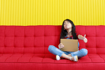 Studio shot of Asian young primary school girl in fashionable denim jeans overalls outfit sitting crossed legs on red couch browsing surfing internet with touchscreen tablet computer in living room