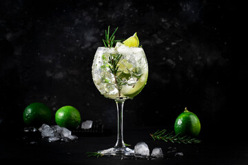Gin tonic alcoholic cocktail drink with dry gin, rosemary, tonic, lime and ice cubes in wine glass. Black bar counter background, bar tools, copy space