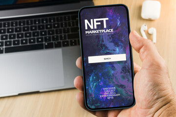 Man holding a smartphone with NFT (Non-Fungible Token) Marketplace app on the wooden table. Office environment