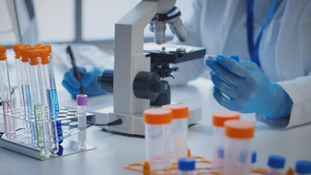 Close up of scientist wearing white coat looking through microscope and making notes whilst holding blood sample in test tube which he puts on desk to show label type a - shot in slow motion