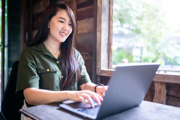 Happy of asian freelance people business female  wearing brownish green dress stylish hipster casual working with laptop computer with coffee cup beside the window of a wooden house background