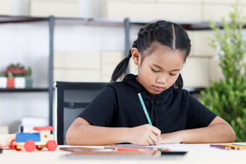 Asian young smart clever elementary schoolgirl artist in casual outfit sitting using color pencils drawing painting picture on paper alone on working table with tablet and wooden toy in living room