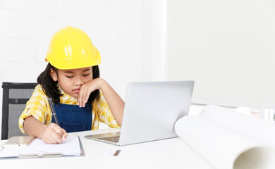 Asian young little girl dreaming about future job career as engineering wearing yellow safety hardhat helmet sitting on working table with laptop computer blueprint smartphone and paper clipboard