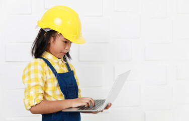 Asian young little preschooler girl future dream job career as engineer and architect wears yellow safety hardhat helmet and overalls standing holding laptop notebook computer in hand typing working