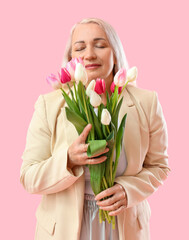 Fashionable mature woman with closed eyes smelling bouquet of tulips on pink background. International Women's Day celebration