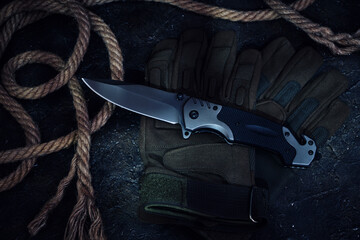 Folding knife, tactical military gloves and rope on dark background.
