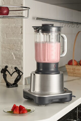A blender with a cooked strawberry smoothie on the kitchen table. Next to a plate with a berry.