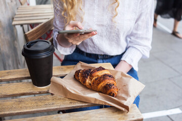 Young white blond woman having a break and holds smartphone in hand. Street veranda table in cafe with cardboard paper cup of coffee to go and french croissant, . business breakfast concept