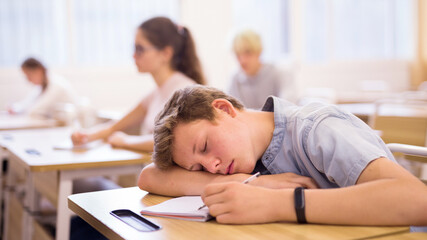 Tired teen student sleeping at desk in classroom during lesson with blurred classmates in background