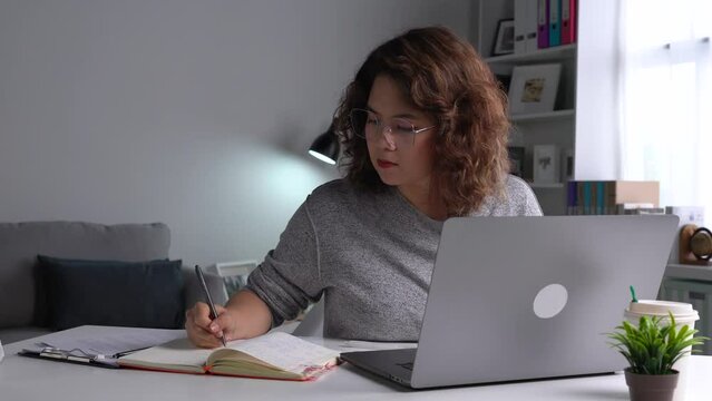 Beautiful Asian woman working on laptop at home. Female student sitting on desk study online on laptop take notes in notebook
