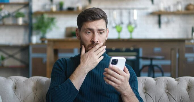 Upset man reacting message on smartphone covers his mouth in fright sitting on sofa at home living room. Unpleasantly Surprised and Scared. Male is shocked by what he saw on display of mobile phone