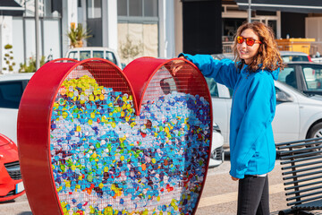 responsible girl hands over the plastic for recycling in a heart-shaped container installed by the authorities on the city street. Awareness and sustainability - obrazy, fototapety, plakaty