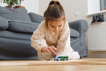 Toddler girl in white dress plays with wooden train at home in the living room 