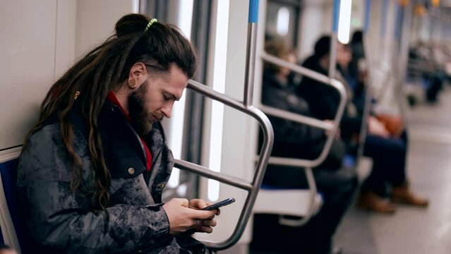 A young man holds a smartphone while riding on a subway train. A bearded man with a phone, touches the touch screen with his fingers.