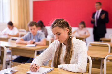 Attractive smart teen girl studying in classroom, listening to lecturer and writing in notebook