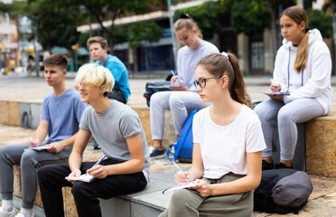 Group of teen students sitting with workbooks in schoolyard in warm autumn day.