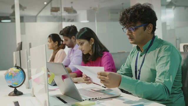 Focused young indian ethnicity businessman working on laptop online sitting at shared table in modern co working open space place with diverse freelance people office lifestyle concept.
