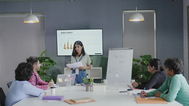 Young Indian female business trainer company manager coach wear suit presenting explain design project result give flip chart presentation training business team group at office workshop meeting.