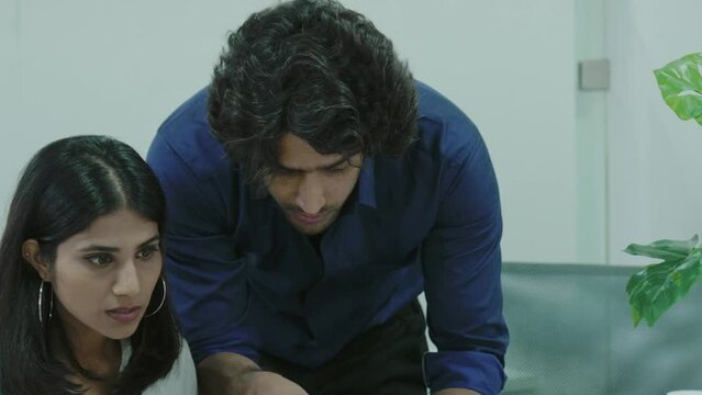 Focused young Asian man project coordinator engaged in paperwork with Indian woman staff member by laptop screen. Skilled male mentor share experience give advice to female apprentice.