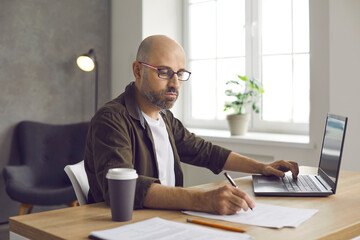 Serious bald business man in glasses sitting at desk in home office, working on laptop computer,...