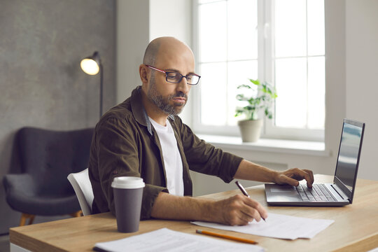 Serious bald business man in glasses sitting at desk in home office, working on laptop computer, searching for information, taking notes, writing down important details, drinking coffee from paper cup