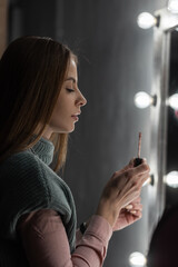 Obraz premium beautiful young girl in fashionable clothes does makeup and paints her lips near a vintage mirror with lights bulbs in the dark