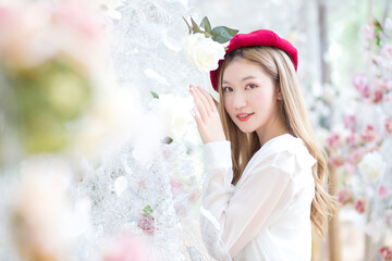 Asian woman has long bronze hair and wears red cap, white dress while she stand looking at camera among flower garden in winter season.