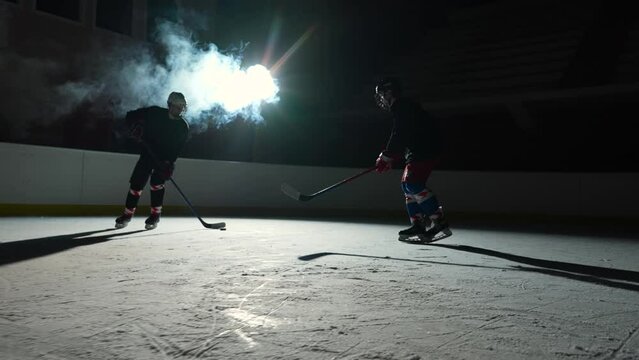 Two men hockey player in uniforms masterfully dribbles, hitting puck with stick and forward scores goal. Hockey puck hits the net. Athletes plays hockey on dark ice arena with spotlights. Slow motion.