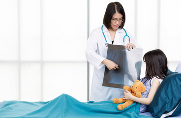 Asian professional friendly female doctor in white lab coat and stethoscope standing smiling showing lungs Xray film to young girl patient lay down on bed hugging teddy bear doll in hospital wardroom