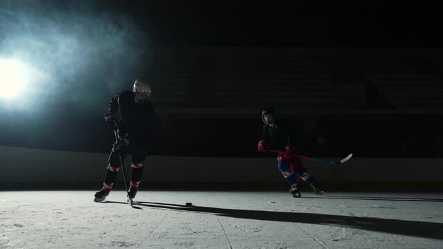 Two sportsmen hockey players who are sliding on ice arena and dribbles, hitting puck with stick. Young guys in uniforms, helmets and with sticks are skating in dark rink with spotlights. Slow motion.