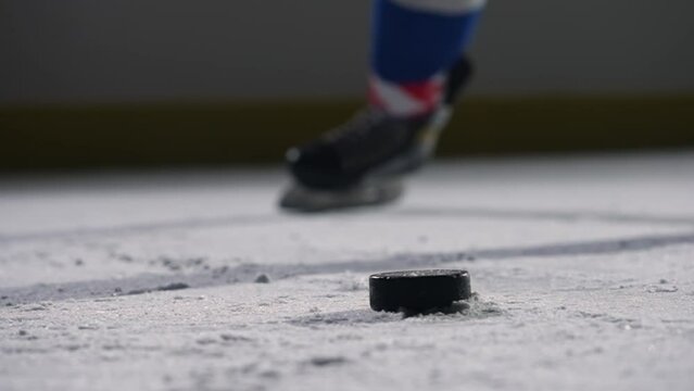 Focus on the black hockey puck lying on the ice of the rink. A hockey player slides across the arena and hits the puck with a hockey stick, cutting the ice into powder. Cinematic slow motion. Close up