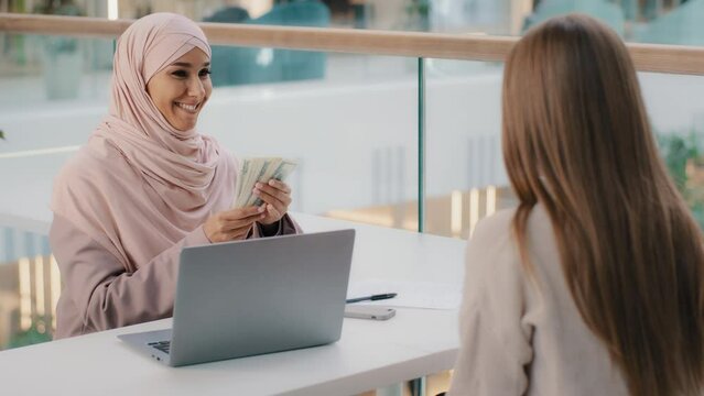 Young friendly smiling arab woman manager sits in office at desk communicates with client explains terms contract gives money unrecognizable caucasian girl withdraws cash from bank account takes loan