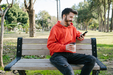 Laughing young man, young man receiving a funny message on phone. Handsome man holding cardboard cup sitting on a park bench.