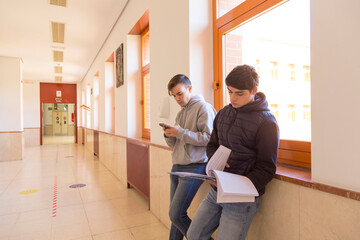 two university student friends in the corridor of the faculty using mobile phones and reading notes