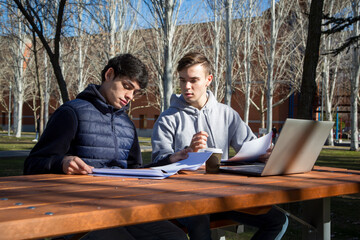 Two young university students studying in a university garden with a laptop computer and papers.