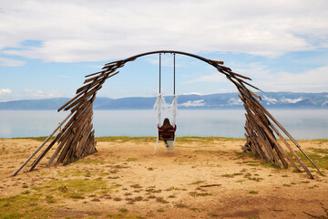 Wooden rustic swing on the beach of Olkhon island. A young girl is sitting on a swing and admiring the beautiful landscape of Lake Baikal. 