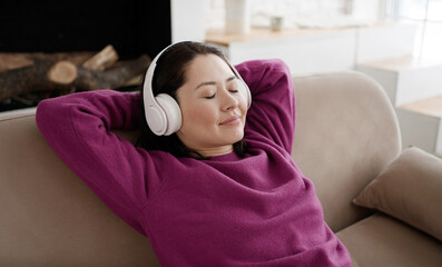 Young asian woman relaxing on comfortable sofa with eyes closed wearing headphones. Pretty mixed race lady enjoys listening chill music audio sound meditating feeling no stress at home.