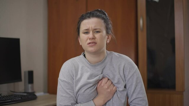 Portrait of young woman with severe pain in chest sitting at home indoors. Sad Caucasian ill lady with heart attack holding chest sighing. Cardiovascular disease concept