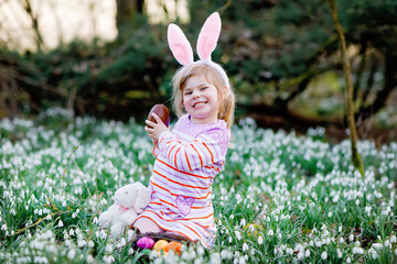 Little girl with Easter bunny ears making egg hunt in spring forest on sunny day, outdoors. Cute happy child with lots of snowdrop flowers, eating huge chocolate egg and colored eggs.