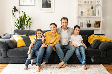 Family garmony. Happy multiracial family of four sitting on the sofa in cozy living room, looking at the camera and smile. Portrait of cheerful spouses and two kids