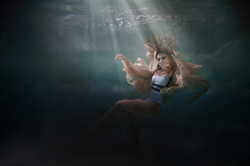 A girl in a light fluttering dress swims underwater as if floating in weightlessness against a dark...