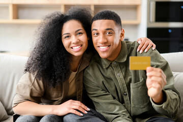 Happy young couple smiling with delight, while sitting at the sofa with credit card and looking at the camera. Happy biracial man holding plastic credit card, preparing to make secure internet payment