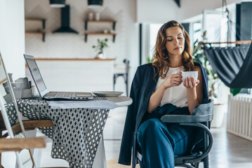 A woman works at home, sitting at a table with a laptop and holding a mug