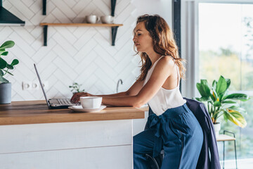 Serious woman sitting at table and typing on laptop in kitchen