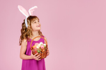 Obraz na płótnie Canvas Cute positive little girl with curly hair in bright dress and bunny ears, smiling happily and looking away from the camera while holding wicker basket with colorful decorated eggs on a pink background