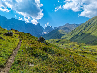 A panoramic view on the sharp mountain peaks of the Chaukhi massif in the Greater Caucasus Mountain Range in Georgia, Kazbegi Region. A hiking trail on a green alpine pasture. Georgian Dolomites.