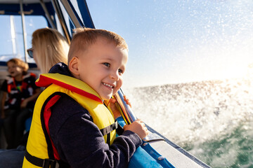 Portrait of cite little blond happy excited smiling caucasian boy wear lifevest enjoy sailing on motor boat sea against blue sky and water splash wave sun backlit. Summer travel vacation recreation - Powered by Adobe