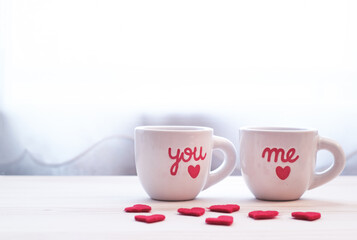 Two valentine's coffee mugs near window with white isolated background, red mini heart on a wooden table. With word you and me on mug. 