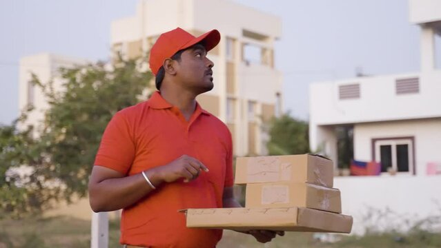 Delivery boy with cardboard boxes in hand searching for address on road - concept of courier service, confused and online shipping